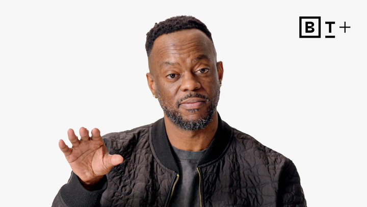 A man wearing a black quilted jacket gestures with his right hand against a plain white background with the B+ logo in the top right corner.