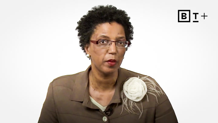 A woman with short curly hair, glasses, and a brown jacket with a white flower brooch sits in front of a plain white background with a "BT+" logo.