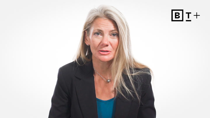 A woman with long blonde hair wearing a black blazer and teal top sits in front of a plain white background with a "BT+" logo in the top right corner.