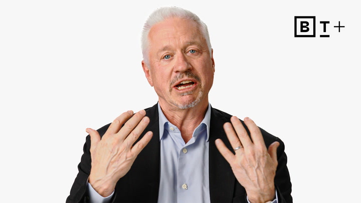 A middle-aged man in a suit gestures with both hands while speaking, against a plain white background with a "B T +" logo in the corner.