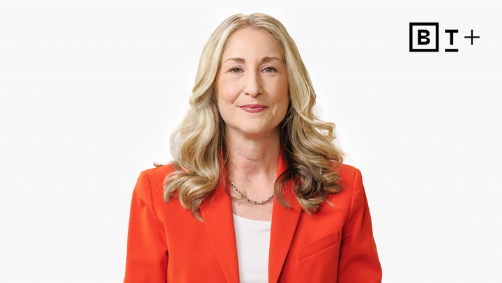 A woman with long blonde hair wearing a bright orange blazer and white top stands in front of a plain white background with a "BT+" logo in the top right corner.