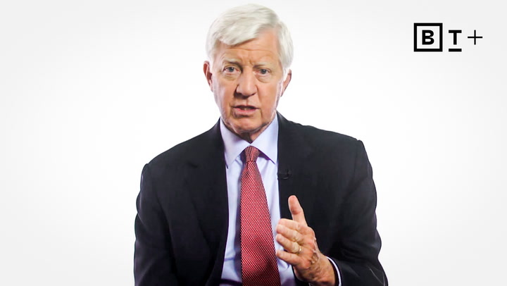 An older man in a dark suit and red tie speaks while gesturing with his hand; a "BT+" logo appears in the upper right corner.