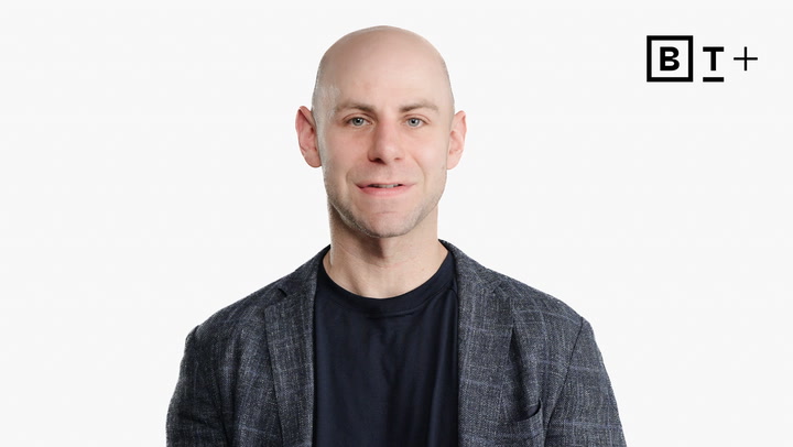 A bald man in a dark blazer and black t-shirt stands against a plain white background with a "B T +" logo in the top right corner.