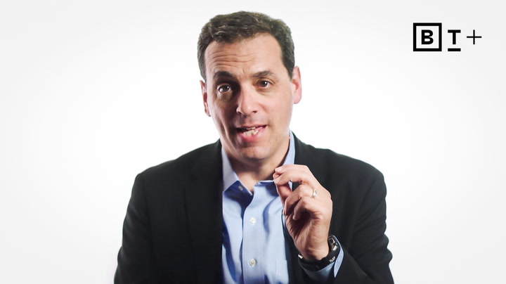 A man in a dark suit and light blue shirt gestures with his right hand against a plain white background with a "B T +" logo in the top right corner.