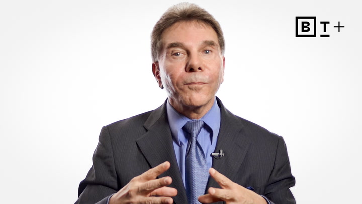 A man in a suit and tie gestures with his hands while speaking against a plain white background with a "B T +" logo in the top right corner.