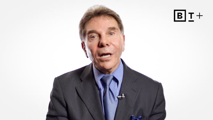A man in a suit and tie speaks in front of a plain white background, with a microphone clipped to his lapel and a "BT+" logo in the top right corner.