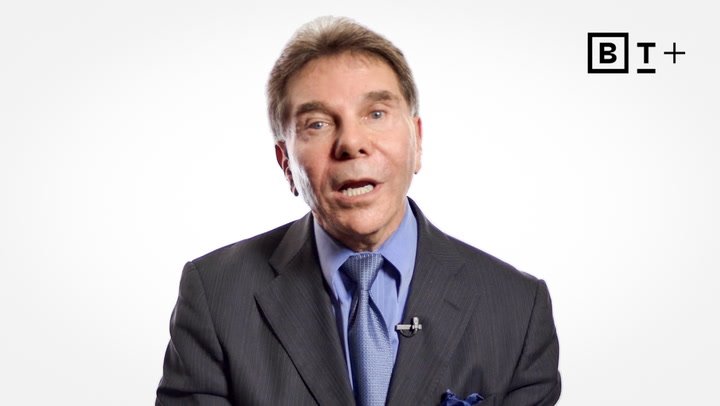 A man in a suit and tie speaks in front of a plain white background, with a microphone clipped to his lapel and a "BT+" logo in the top right corner.