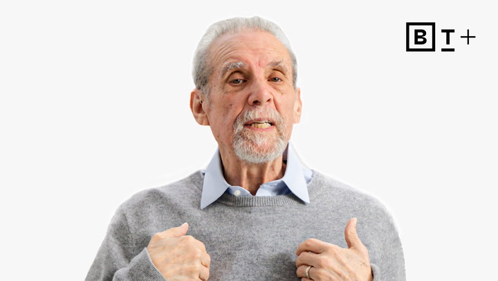 An older man with gray hair and beard, wearing a gray sweater over a light blue shirt, gestures with both hands; a "B T +" logo appears in the top right corner.