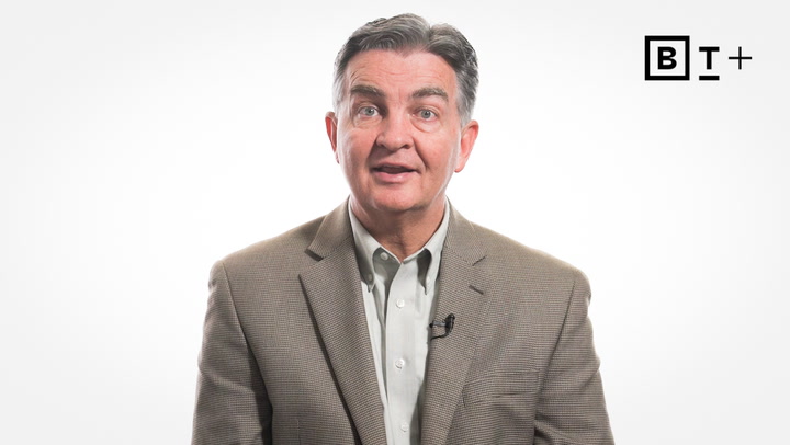 A man in a brown blazer and light shirt is speaking in front of a plain white background with a "BT+" logo in the top right corner.