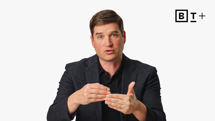 A man in a dark blazer and black shirt gestures with his hands while speaking, against a plain white background with a "B T +" logo in the top right corner.