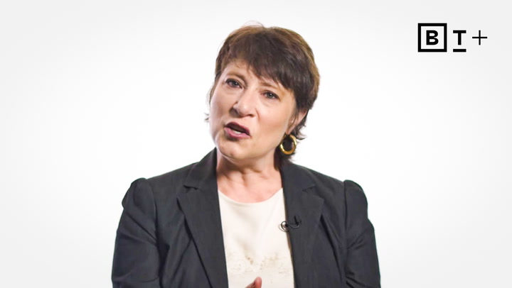A woman with short brown hair in a black blazer and white top speaks in front of a white background with a "B T +" logo in the top right corner.