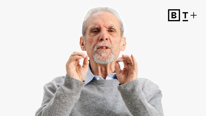 An older man with gray hair and a beard gestures with both hands while speaking, wearing a gray sweater over a blue shirt. A black and white "B T +" logo appears in the top right corner.