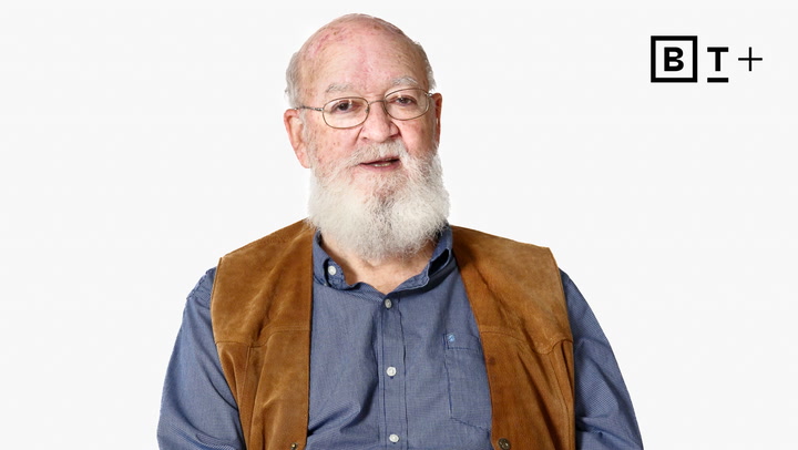 An older man with a white beard and glasses wears a blue shirt and brown vest, seated against a plain white background. A "B T +" logo appears in the top right corner.