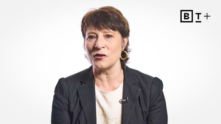 A woman with short brown hair wearing a black blazer and white blouse speaks in front of a white background with a microphone clipped to her lapel. A black "B T +" logo is in the top right corner.