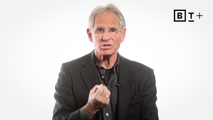 An older man in a dark blazer gestures with his fist raised in front of a white background with a "BT+" logo in the top right corner.