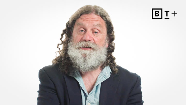 A man with long curly hair and a gray beard wearing a blazer and open-collar shirt sits in front of a white background with a "BT+" logo in the top right corner.