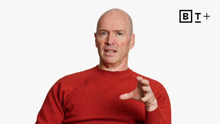 A bald man in a red sweater gestures with his right hand against a plain white background with the BT+ logo in the top right corner.