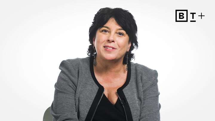 A woman with short dark hair wearing a gray blazer and black top sits and smiles in front of a plain white background with a "BT+" logo in the top right corner.