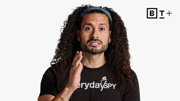 A man with long curly hair wearing a black "Everyday Spy" t-shirt gestures with his right hand; a BT+ logo appears in the top right corner against a plain white background.