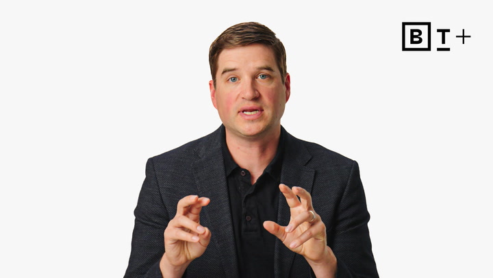 A man in a dark blazer and black shirt speaks while making air quotes with his hands, with a "BT+" logo in the top right corner on a plain white background.