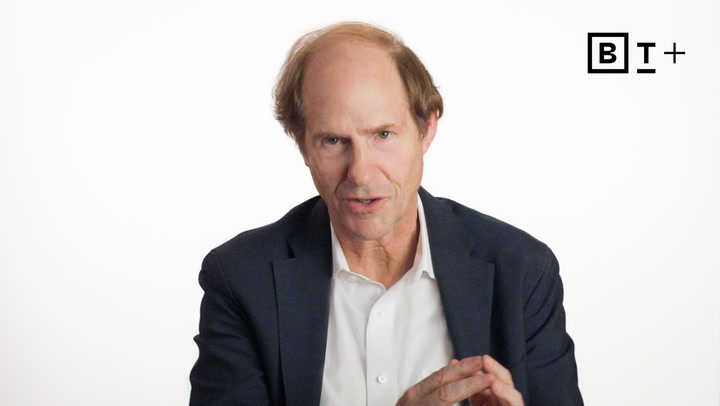 A middle-aged man in a suit jacket sits against a plain white background with a “B T+” logo in the top right corner.