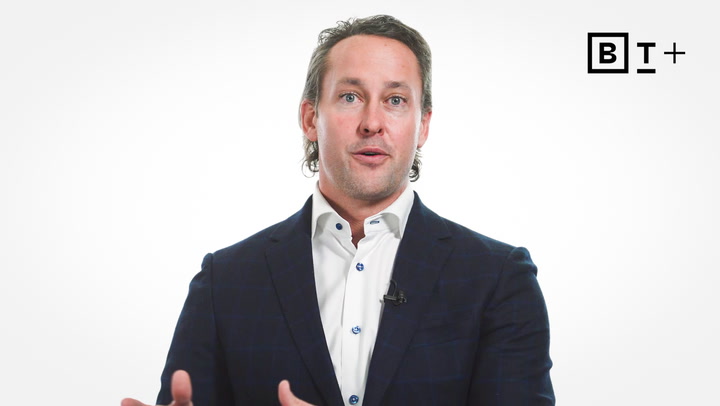 A man in a dark blazer and white shirt speaks while gesturing, with a "BT+" logo in the top right corner on a plain white background.