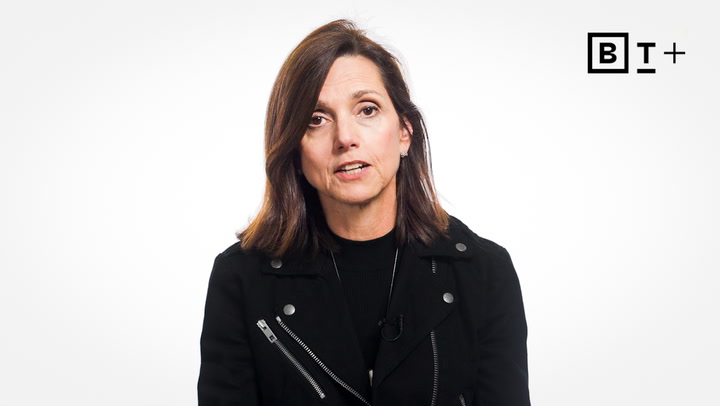 A woman with shoulder-length brown hair wearing a black jacket sits in front of a white background with a "B T +" logo in the top right corner.