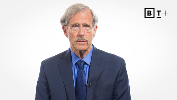 An older man in a suit and tie sits against a plain white background with a "BT+" logo in the top right corner.