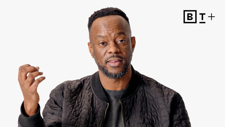 A man with a short beard and short hair, wearing a black textured jacket, gestures with his left hand against a plain white background with the "B T +" logo in the corner.