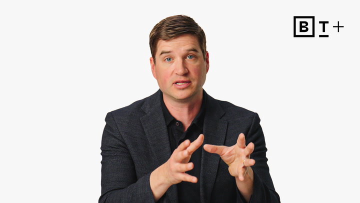 A man in a dark blazer gestures with his hands while speaking in front of a plain white background with a "BT+" logo in the top right corner.