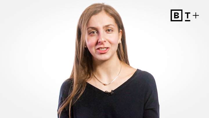 A woman with long brown hair, wearing a black top and necklace, speaks in front of a white background with a "BT+" logo in the top right corner.