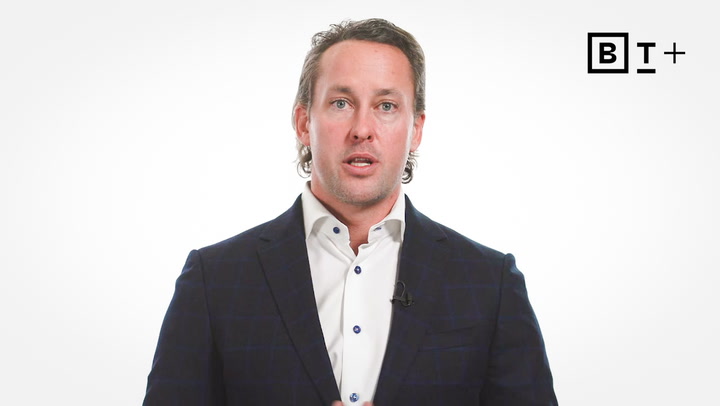 A man in a dark suit and white shirt speaks in front of a plain white background with the B T Plus logo in the top right corner.
