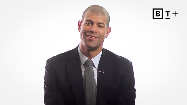 A man in a suit and tie sits and smiles in front of a plain white background with a "B T +" logo in the top right corner.