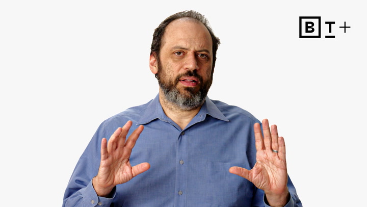 A man with a beard, wearing a blue shirt, gestures with both hands in front of a plain white background with the "B T +" logo in the top right corner.