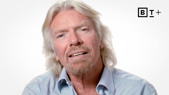 Man with long blond hair and a light beard wearing a light blue shirt, sitting against a plain white background with a "B T +" logo in the top right corner.