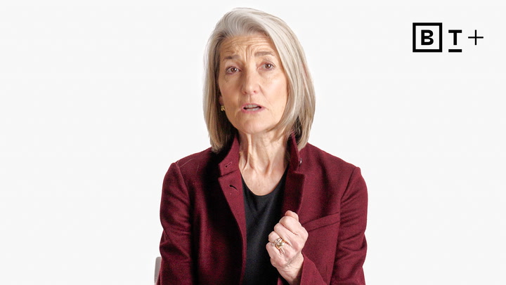 A woman with gray hair wearing a maroon blazer sits against a white background, speaking and gesturing with one hand. The BT+ logo is visible in the top right corner.
