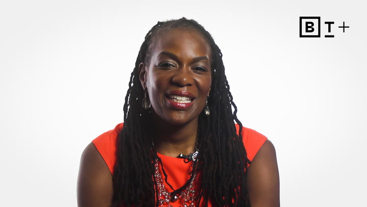 A woman with long braids wearing a bright orange top and necklace smiles at the camera, with a "B T +" logo in the top right corner against a white background.