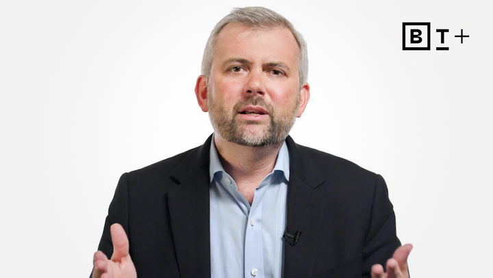 A man in a suit gestures with his hands while speaking in front of a white background with a "B T +" logo in the top right corner.