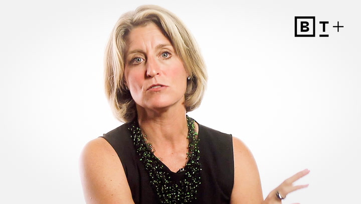 A woman with short blonde hair wearing a sleeveless black top and beaded necklace speaks against a plain white background. A "BT+" logo appears in the top right corner.