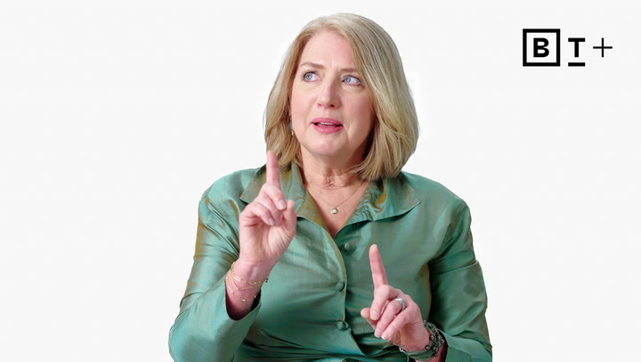 A woman with shoulder-length blonde hair wearing a teal blouse gestures with both index fingers raised, seated against a plain white background with a "B T +" logo in the corner.