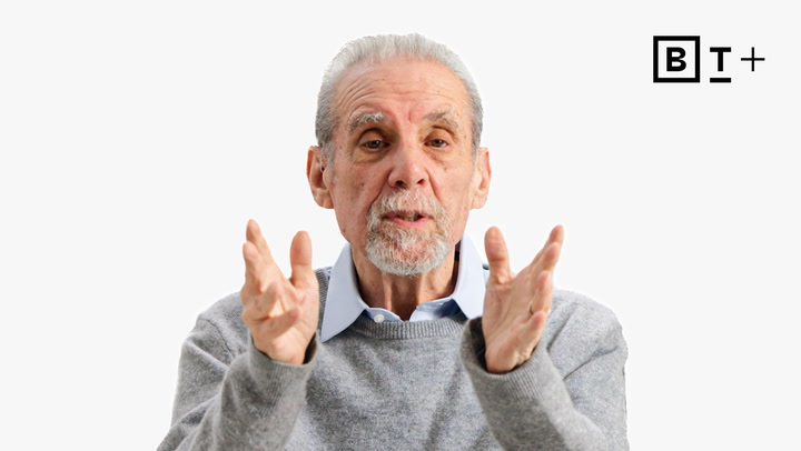 An older man with gray hair and a goatee speaks while gesturing with both hands. He wears a gray sweater over a collared shirt, with a "B T +" logo in the top right corner.