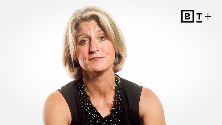 A woman with short blonde hair and a neutral expression faces the camera. She wears a sleeveless black top and beaded necklace. The background is plain white with a "BT+" logo in the top right corner.