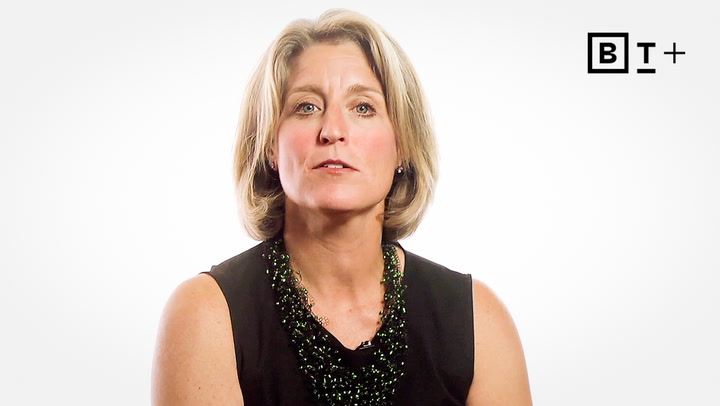 A woman with short blonde hair wearing a black sleeveless top and green beaded necklace sits in front of a plain white background with a "BT+" logo in the top right corner.