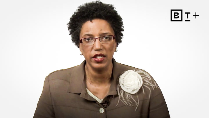 A woman with short curly hair, glasses, and a brown blazer with a large white flower brooch speaks in front of a plain white background with a BT+ logo in the top right corner.