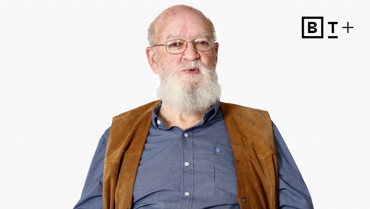 An older man with a white beard and glasses wears a blue shirt and brown vest, seated against a plain white background with a "B T +" logo in the upper right corner.