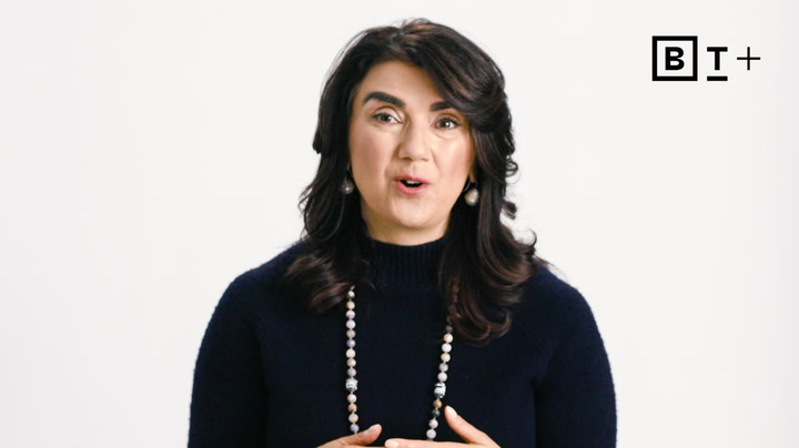 A woman with dark hair wearing a dark sweater and beaded necklace speaks in front of a plain white background with a “BT+” logo in the top right corner.