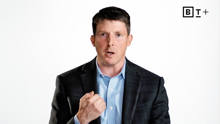A man in a dark suit and light blue shirt speaks while gesturing with his hand. A "BT+" logo appears in the top right corner against a plain white background.