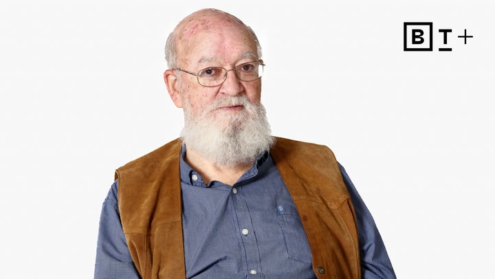 An older man with a white beard, glasses, and a brown vest over a blue shirt sits against a plain white background with a "B T +" logo in the top right corner.