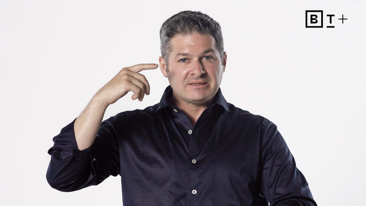 A man in a dark shirt points to his head with his right hand. The background is plain white, and the letters "BT+" are in the top right corner.