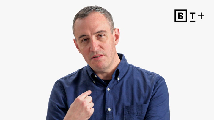 A man in a blue button-up shirt gestures with his right hand against a plain white background with a "B T+" logo in the top right corner.